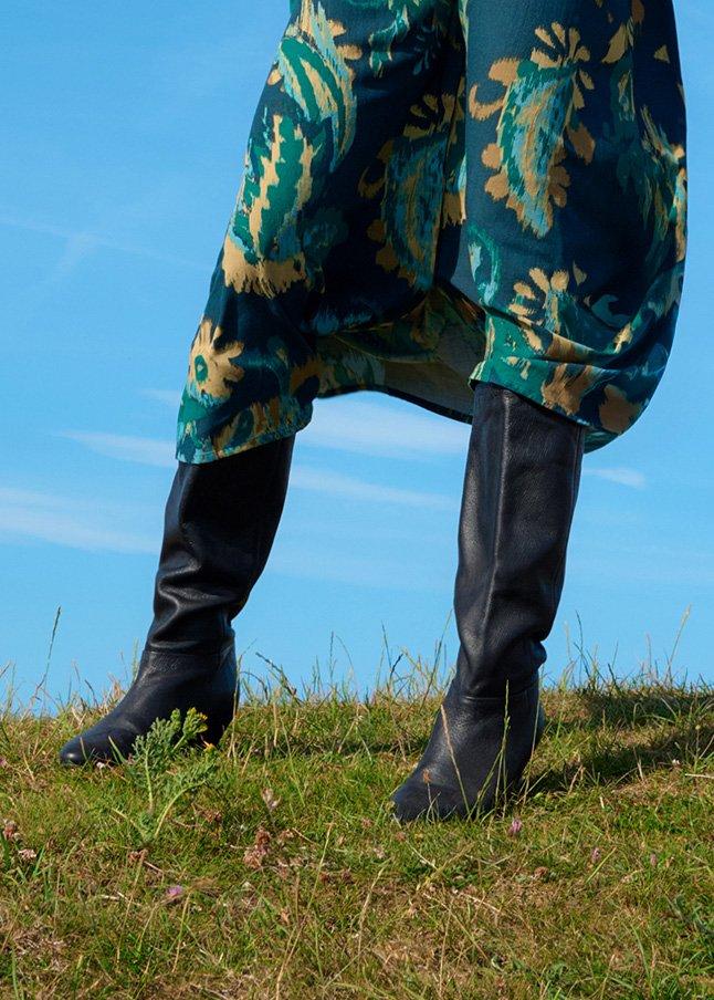 a woman in a green dress and black boots standing on a grassy hill